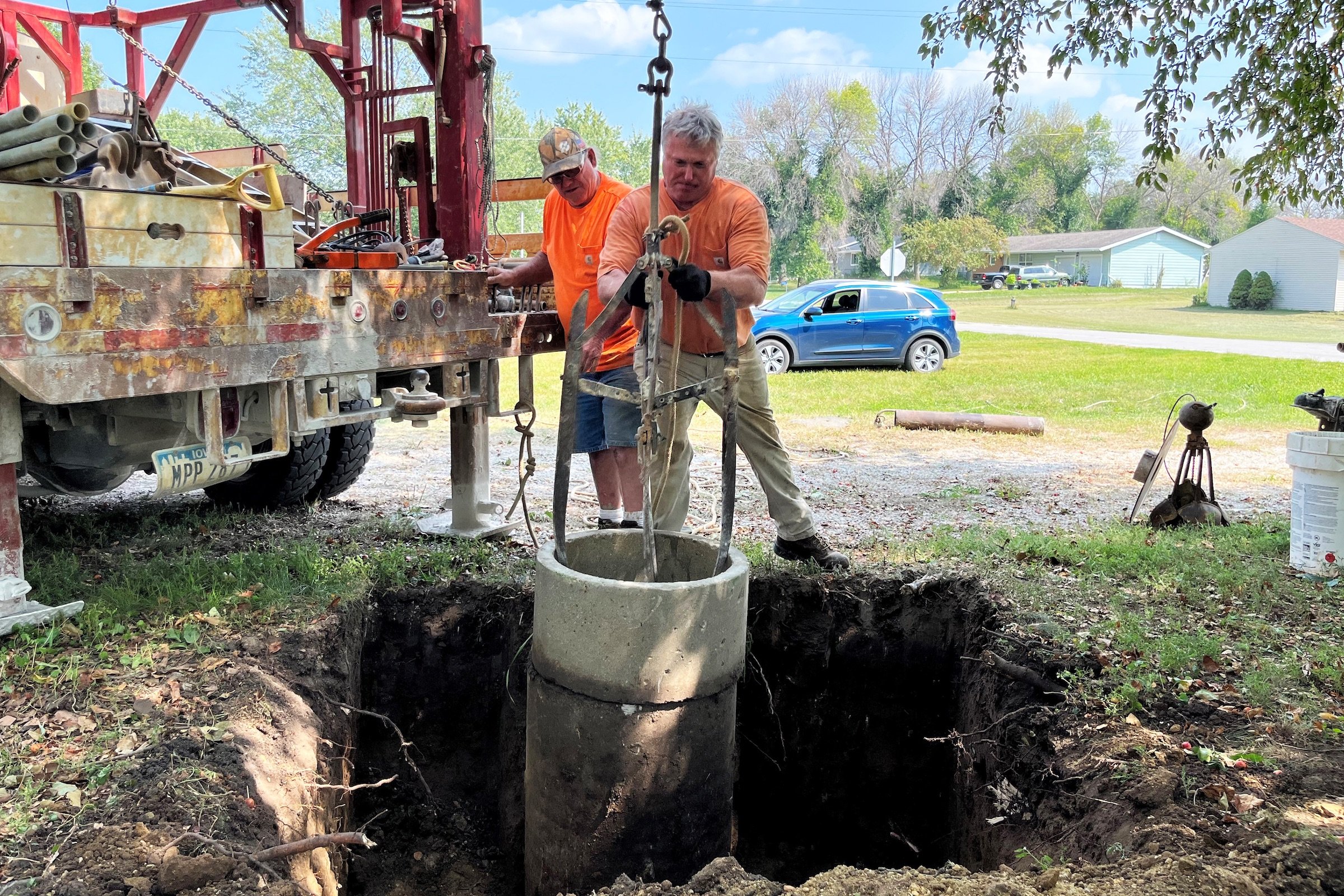 Two men in orange shirts using a machine to lower a concrete cylinder into a hole in the ground.
