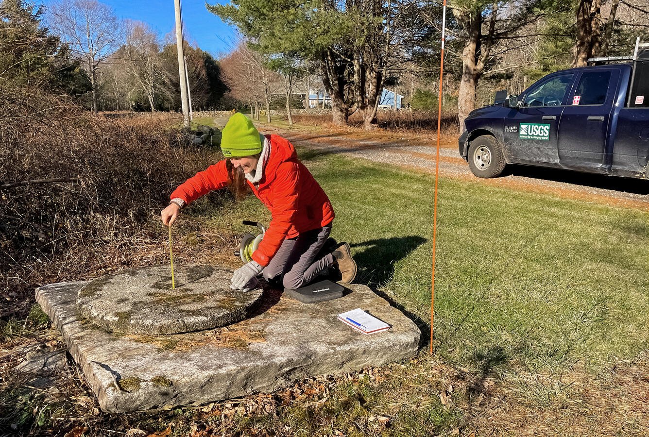 A person in a red jacket kneeling beside a concrete structure taking water level measurements.
