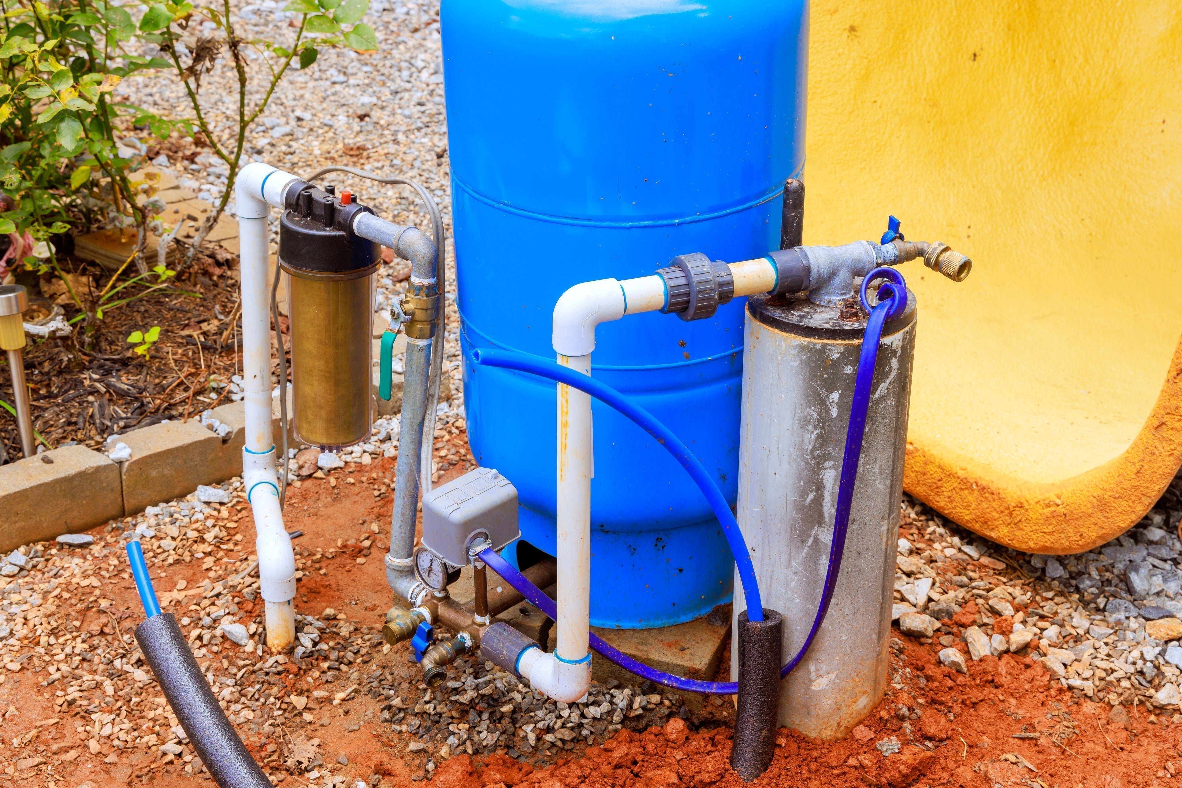 A blue water pump system with various pipes and a pressure gauge is installed on a gravel and dirt surface, near some plants.