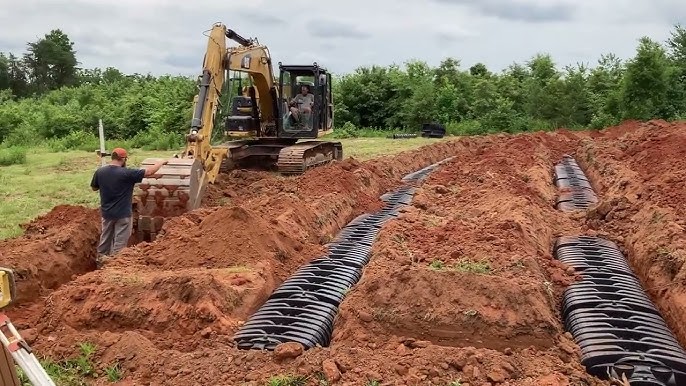 Construction site with a person overseeing a trench being dug by an excavator, with plastic drainage pipes laid in parallel trenches.