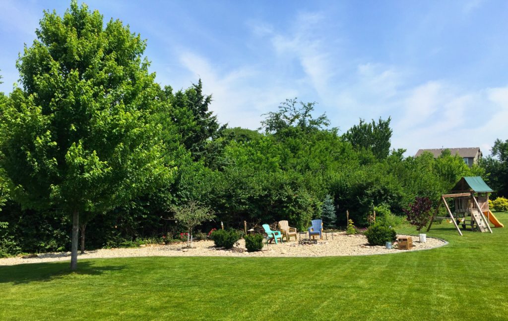 A backyard with a well-maintained lawn, a variety of trees and bushes, a gravel sitting area with several chairs, and a children's playset with a slide.