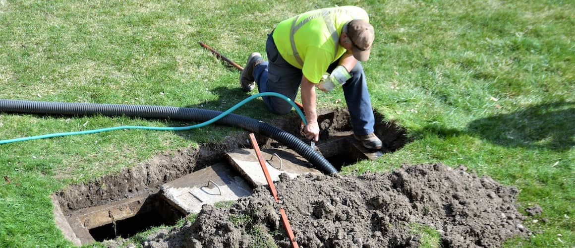 A worker in a yellow shirt and cap is kneeling on the grass, handling a large hose near an open septic tank.