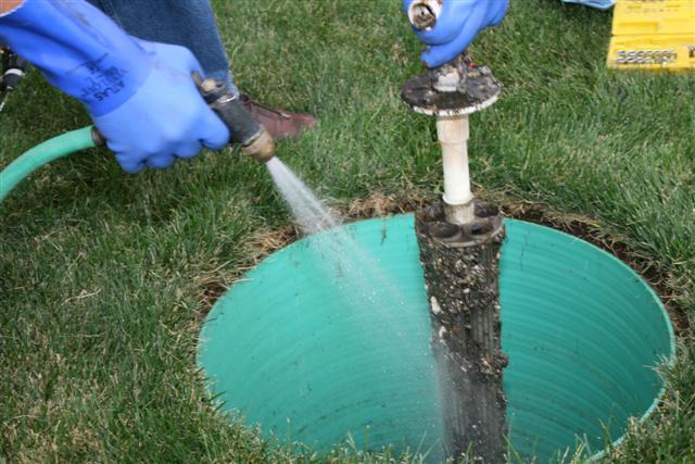 A person wearing blue gloves uses a hose to clean a septic system riser in a grassy area.