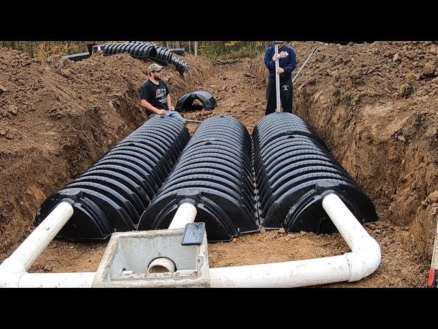 Two people are installing a septic system in a dug-out area with black, dome-shaped infiltration chambers connected by white pipes.