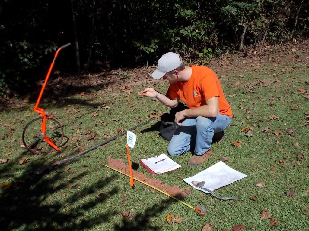 A person in an orange shirt and cap is kneeling on the grass beside some measurement and survey equipment, closely examining something in their hand.