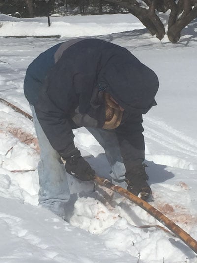 A person wearing a hooded jacket and gloves bends over while handling a long wooden stick in a snowy landscape.