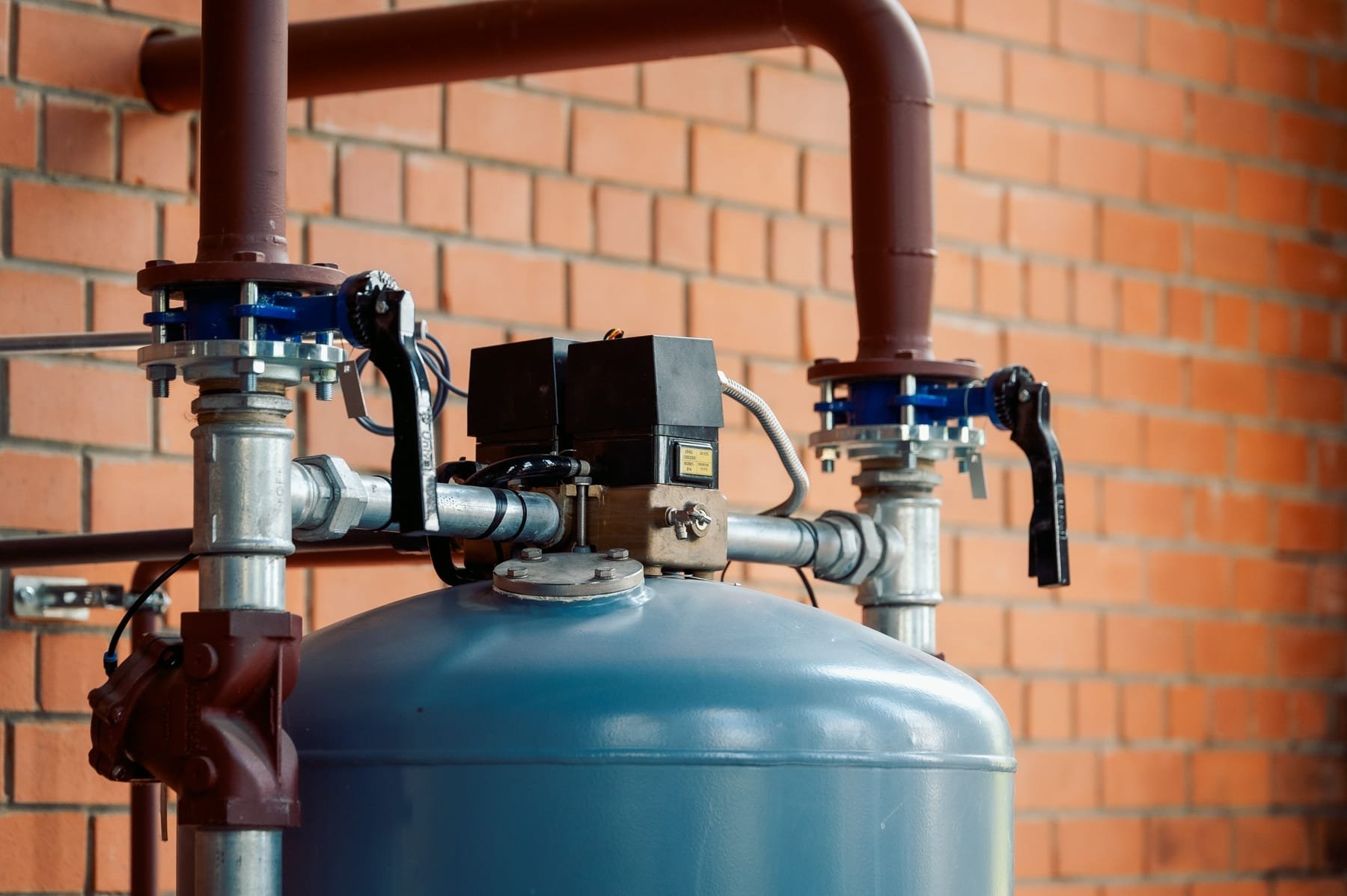 Close-up of industrial metal pipes, valves, and a pressure tank against a brick wall background.