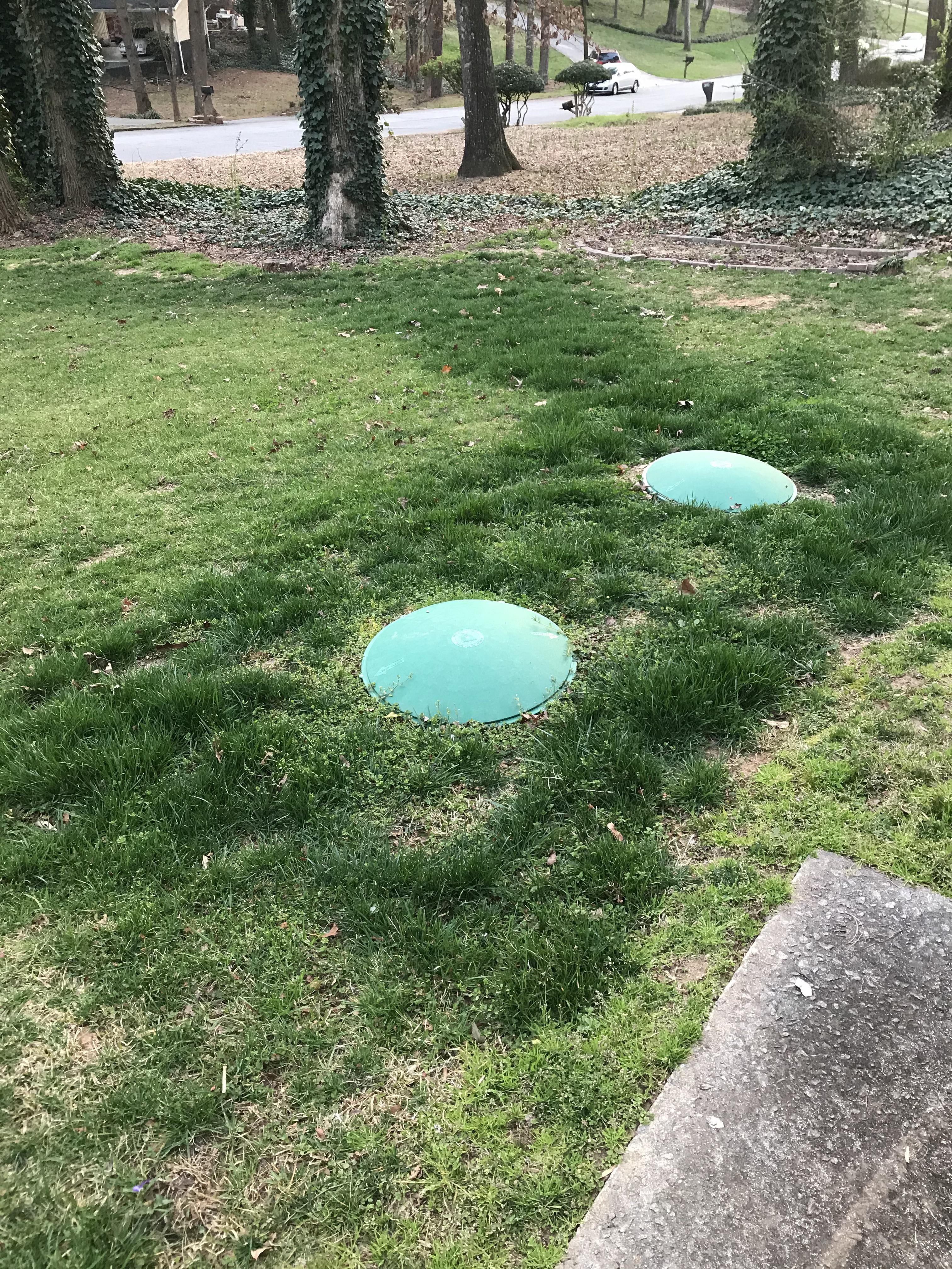 Two green manhole covers on a grassy lawn with trees and a street in the background.
