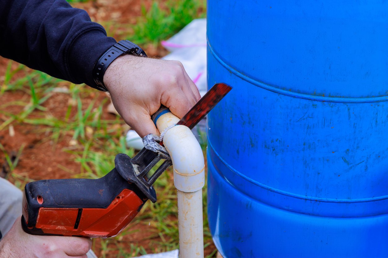 A person uses a reciprocating saw to cut a white plastic pipe beside a large blue barrel.